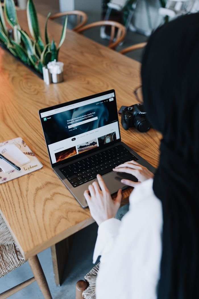 Woman in hijab working on a laptop at a wooden table, camera nearby.