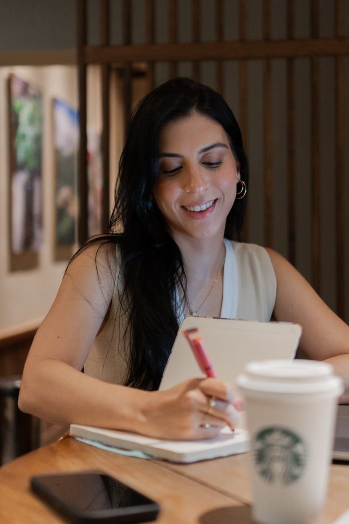 Smiling woman writing notebook at café table with coffee.