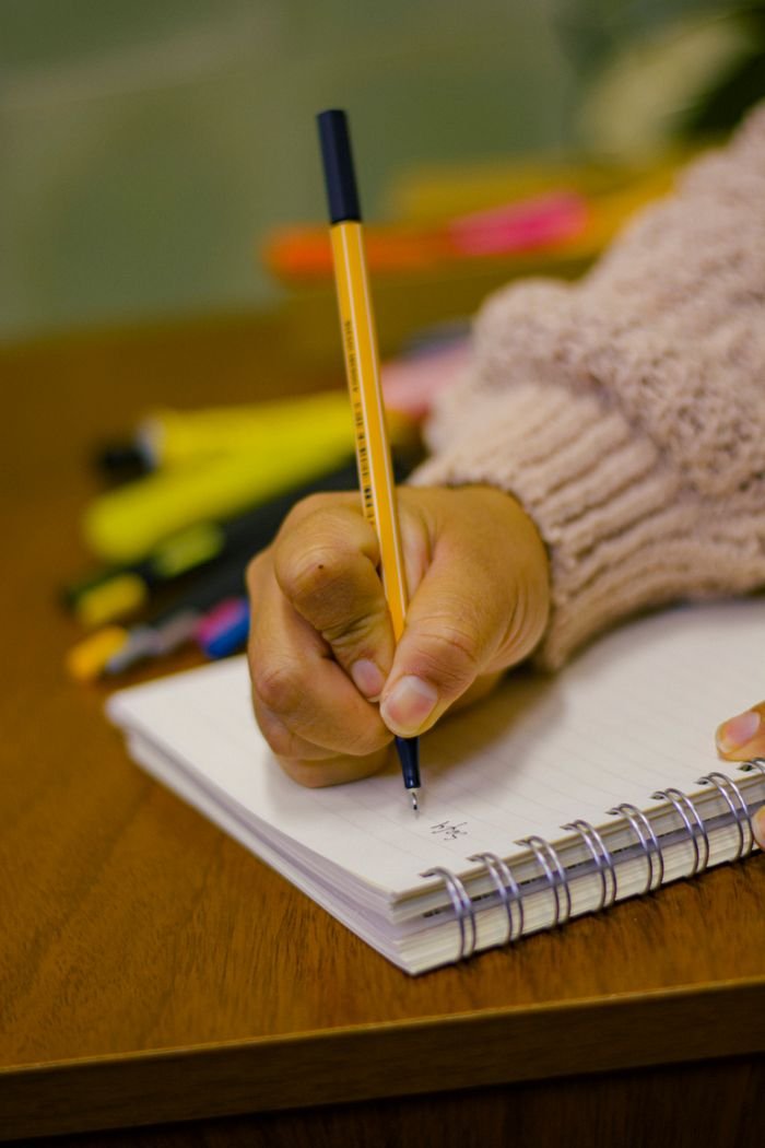 A person's hand writing in a spiral notebook using a pen, surrounded by colorful pens.