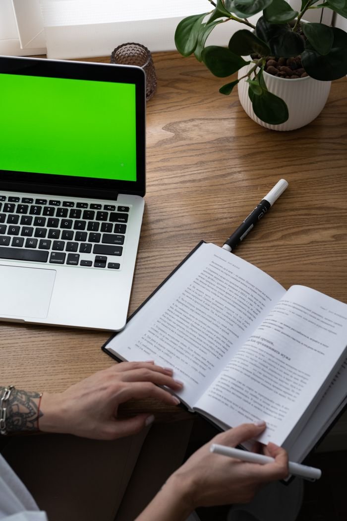 High angle of crop unrecognizable tattooed female student doing homework assignment sitting at wooden table with textbook and laptop
